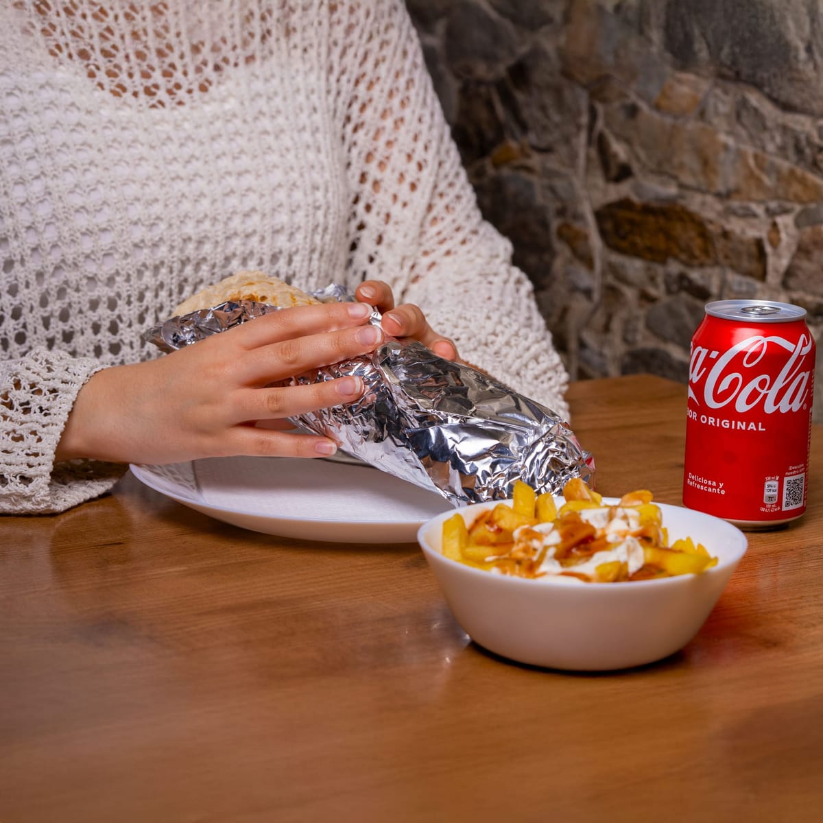 Person isst an einem Holztisch ein eingepacktes Sandwich mit Pommes und einem Softdrink